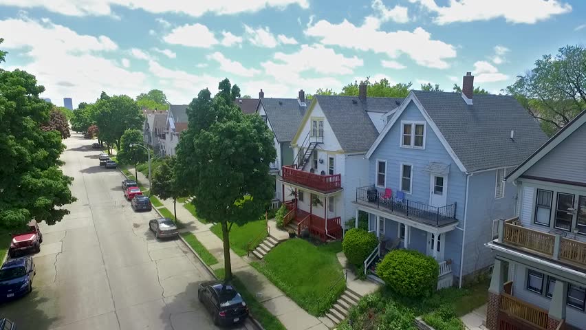 An aerial drone shot of a Milwaukee neighborhood showing streets, cars, trees, houses, blue sky, clouds and the city in the background on a bright and sunny day.