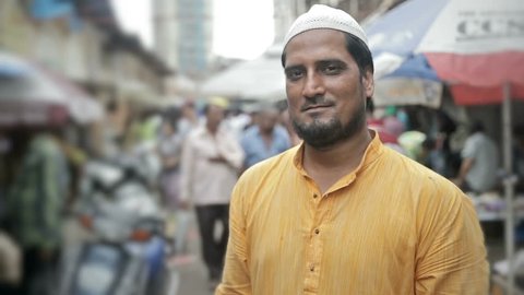 A mid shot of a confident smiling Muslim young man standing and looking into a camera in a busy market