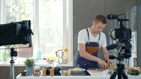 Male chef in apron standing at kitchen countertop and sprinkling spring onions on hot scrambled eggs, then adding cherry tomatoes and talking before digital camera  while recording cooking show