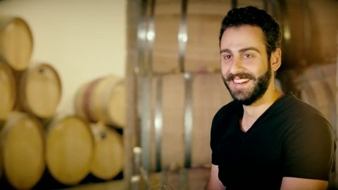 Man sommelier sniffing aroma red wine in glass on background old cellar with barrels. Close up bearded winemaker tasting beverage on wine factory