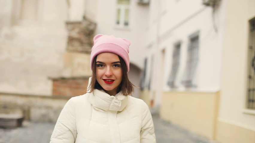 Brunette pretty young women portrait in cute pink hat and red lips smiling, flirting and looking in to the camera. Old city background. Cold season. Slow motion.
