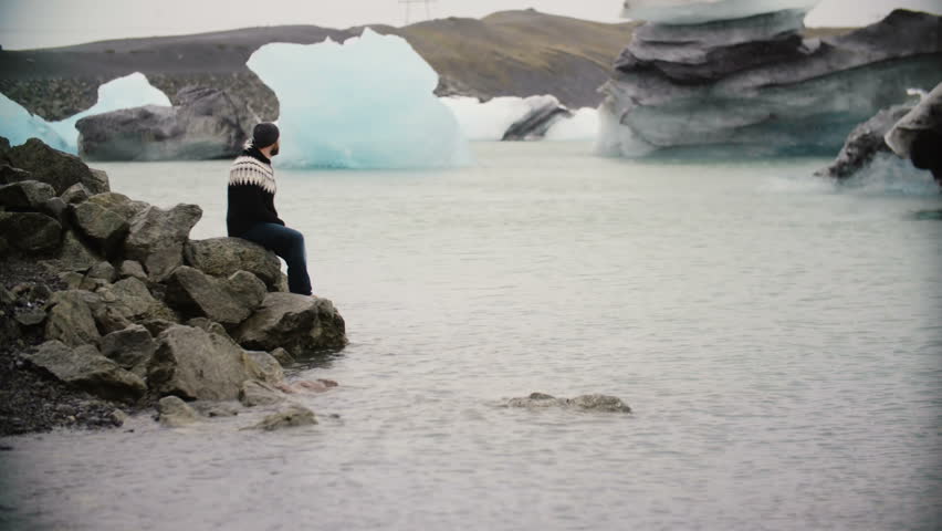 Young handsome man sitting on the rock in ice lagoon and enjoying the view. Male exploring the Iceland alone.