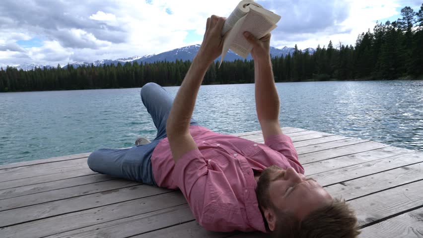 Young caucasian male lying down on wooden pier above mountain lake reading a book to relax and enjoy nature. People enjoying freedom and relaxation moments concept. Shot in Jasper national park, Canad