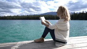 Young caucasian female lying down on wooden pier above mountain lake reading a book to relax and enjoy nature. People enjoying freedom and relaxation moments concept. Shot in Jasper national park, Can