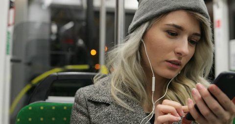Portrait of cute girl in headphones listening to music and browsing on mobile phone in public transport. City lights background