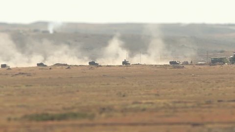 A military convoy of tanks and armored vehicles rides through the desert raising dust. Moving military equipment in the sand against the background of mountains.
