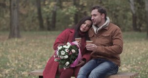 Loving couple sit on bench drink hot coffee tea in autumn forest park 4k date video. Young man and overweight fat woman with flowers speaking. Love story romance concept