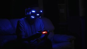 A man plays computer games in a helmet of virtual reality. The helmet and the joystick are lit. It's dark in the room