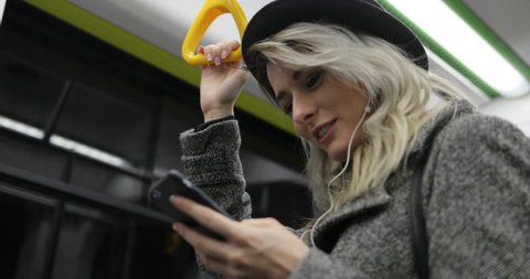 Portrait of cute girl in headphones holds the handrail, listening to music and browsing on mobile phone in public transport. City lights background
