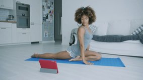 Beautiful confident woman in shorts stretching body on mat while watching tutorial video with yoga practice using tablet.