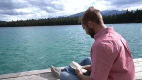 Young caucasian male lying down on wooden pier above mountain lake reading a book to relax and enjoy nature. People enjoying freedom and relaxation moments concept. Shot in Jasper national park, Canad