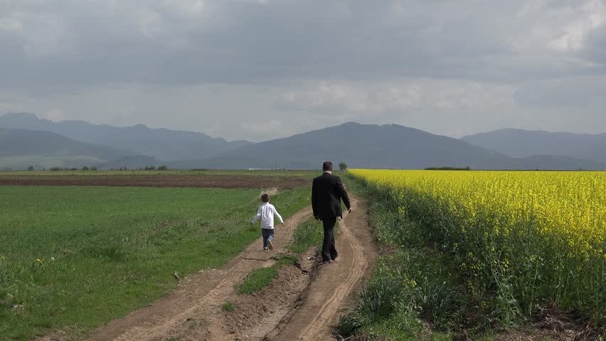 Businessman and his son with back walking on countryside path, yellow rape field, father and child walking next to each other on alley