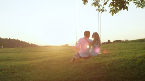SLOW MOTION COPY SPACE: Embraced girlfriend and boyfriend on a tree swing kiss in sunset filled countryside. Couple bonds on a summer date in nature, hugging and sharing a kiss on a swaying rope swing