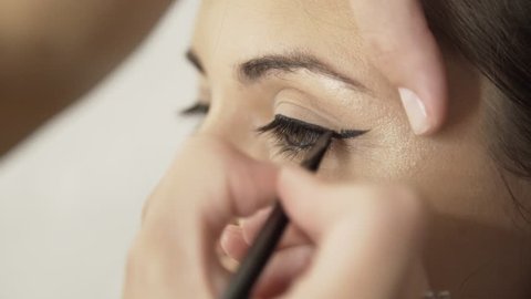 Hand of an unrecognizable make up artist applying black eyeliner to an eye of a girl with fair hair. A side view. Handheld real time medium shot