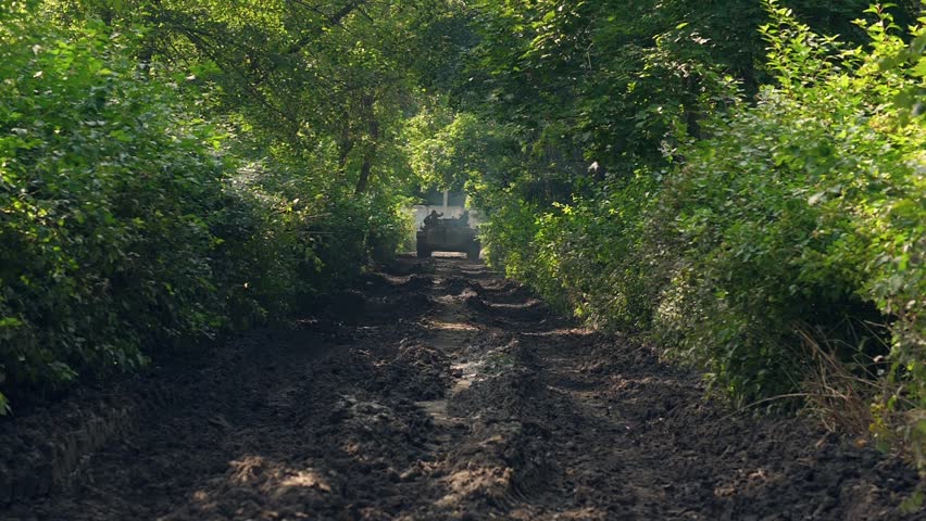 Military soldiers sitting on moving war tank on road through green forest back view. Army tank with soldiers driving on military training slow motion