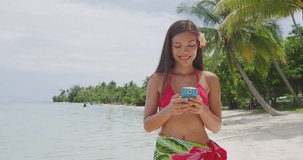 Woman using smart phone on beach - Bikini travel girl using smarphone on holiday smiling happy on Matira Beach, Bora Bora. Woman on holidays beach vacation wearing pareo in Tahiti, French Polynesia.