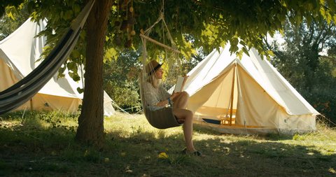 Beautiful Young Woman Reads Book while Sitting in the Hammock Chair under the Tree. Girl Wears a Hat, in the Idyllic Background with Green Trees and Camping Tents. Shot on RED Epic 4K Camera.