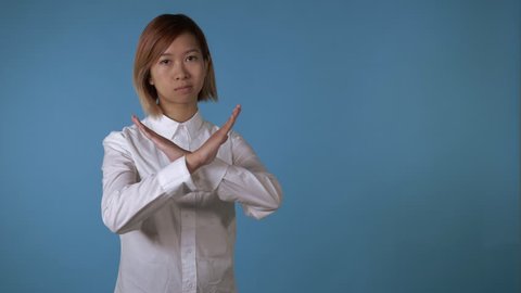 portrait young asian female posing showing hand gesture stopping crossing harms on blue background in studio. attractive korean woman with blond hair wearing white casual shirt looking at the camera