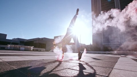 Young woman with colorful smoke grenade jumping high performing a flip outdoors in city. Female with smoke sticks practicing tricking over urban city space.