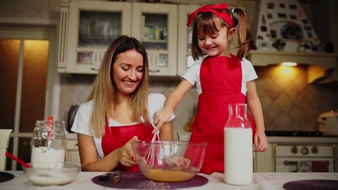 Beautiful young mother helping her little daughter along to cook cake in red aprons. Pour the flour into a bowl and beat the batter to make a cake in the kitchen.