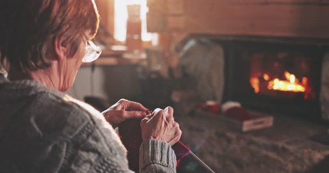 Senior woman hands knitting by the fireplace. Slow motion. Unrecognisable grandmother relaxes by warm fire making handmade gifts for her family. Cozy atmosphere. Winter and Christmas holidays concept. 스톡 비디오