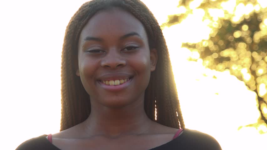 Young beautiful black woman smiles to camera in park during sunset with sun rays