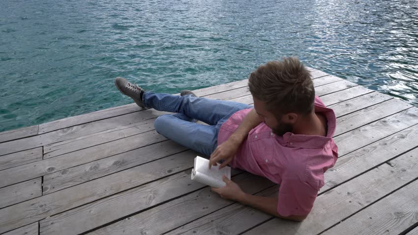 Young caucasian male lying down on wooden pier above mountain lake reading a book to relax and enjoy nature. People enjoying freedom and relaxation moments concept. Shot in Jasper national park, Canad
