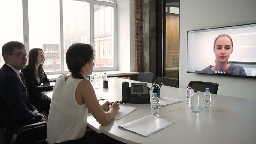 Young employees attend online conference in modern office. Man in glasses, surrounded by two women, looks attentively at TV screen hanging on wall, where they see female coach who has contacted them | Shutterstock HD Video #33565306