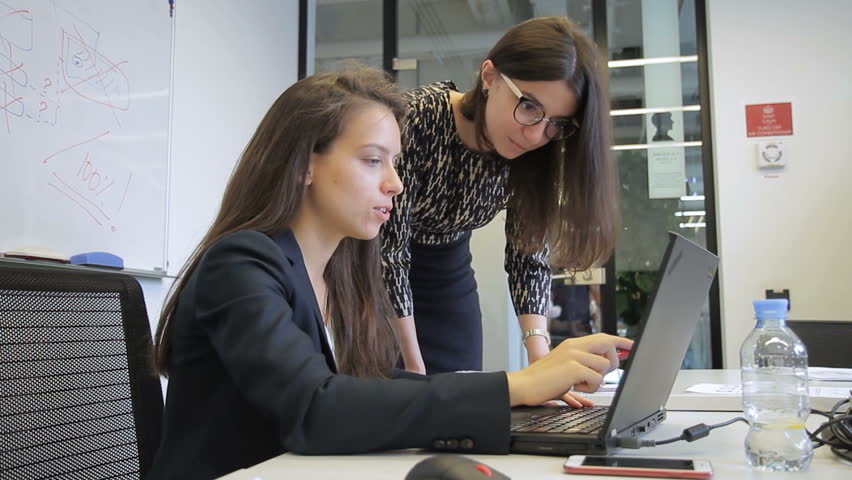 Young women are talking, looking at PC screen in modern office. Brunette demonstrates to colleague information on display of black laptop, pointing with pen then gives out paper documents for making Royalty-Free Stock Footage #33565669