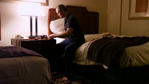 A man gets ready for bed in a hotel room and turns off the light before lying down in his bed