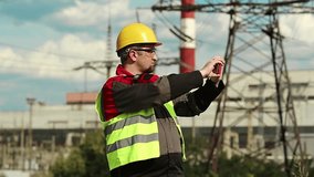 Powerman records videos on red smartphone. Workman in yellow hard hat with smartphone at electric power plant