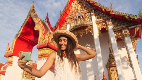 Attractive Young Mixed Race Tourist Girl in White Dress and Big Straw Hat Taking Selfie Photo with Mobile Phone at Thai Buddhist Temple. Phuket, Thailand. 4K, Slowmotion.
