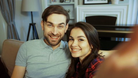 Close up of young charming brunette woman and attractive man sitting on the sofa and making selfies in the room at home. POV. Indoors. Portrait shot