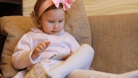 A little cute girl sitting in sofa in a living room, looking at the phone screen cartoon and playing the game. Funny child plays downloaded application on a smartphone