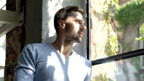 Handsome man in blue sweater sitting on window's sill and enjoys sunlight, steadycam shot
