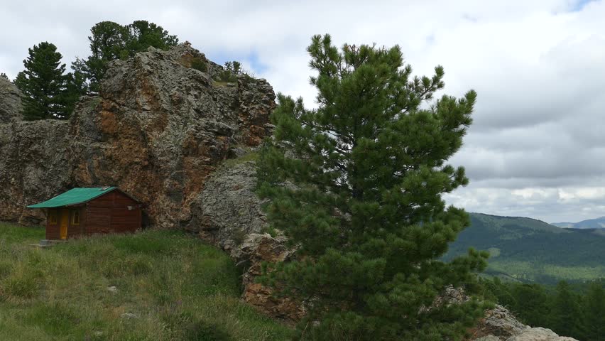 A small pine and a hut on top of a cliff against mountains and a cloudy sky on a summer day.