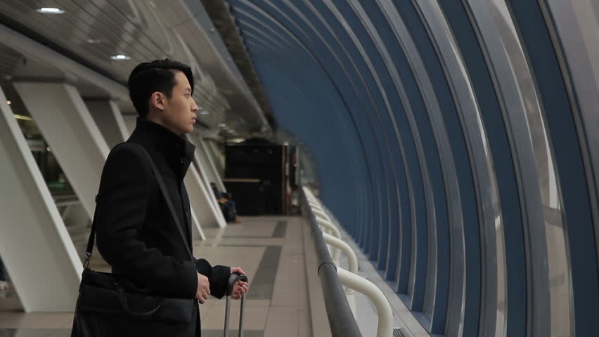 Handsome korean man is walking along airport building indoors. Guy stands with heavy suitcase in hand and looks ahead, then going confidently. 