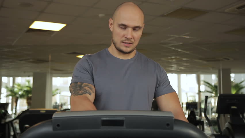 The portrait of handsome spotrsman who enjoys running on the race track in modern gum. The professional turns on the equipment and starts his exercises. The man with tattoo on his shoulder wears gray
