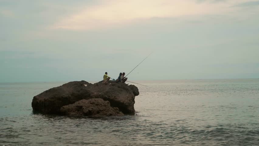  Fishermen sit on a large stone in the sea and fish on fishing rods. Three men fishing on a rock in the water. Sunset, Clouds, waves. Friends fishing, summer day. Slow motion