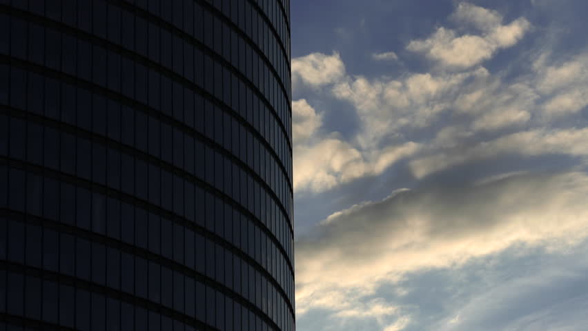 Timelapse with evening clouds flying past steel and glass round-shaped skyscraper corporate office building. (av40184c)