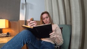 in the evening, the woman sits on a comfortable armchair in the living room, talks on video from her smartphone and makes notes in a notebook. 4k