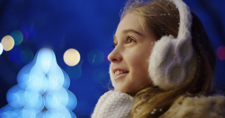 Close up of the happy cute girl looking charmed at something and clapping in white winter gloves on the blurred Christmas lights in the night. Portrait shot. Outside