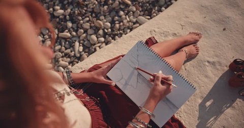 Young woman in boho style drawing nature scene in sketchbook at the beach at sunset