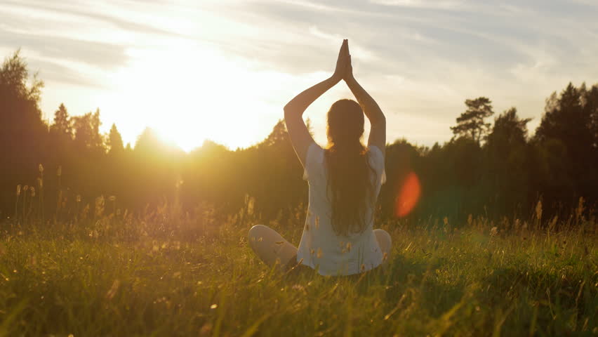 The view of young female from behind sitting in field. Relaxed healthy woman breathing deep, meditating and practicing yoga in peace and harmony with nature at sunset with lens flare. | Shutterstock HD Video #33635581