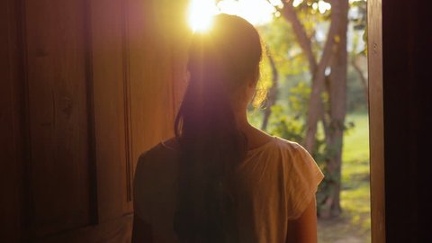 Young woman opening the door and slowly walking outside. Female escaping the city life and enjoying nature and freedom, breathing fresh air outdoors early in the morning. Lens flare.