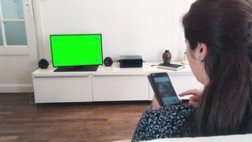 Dolly Shot Millennial Woman Using Smartphone Watching Television. Charming young woman using a smartphone and watching green screen television. Shot behind models shoulders.