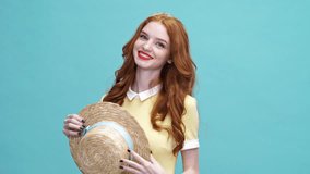 Happy ginger woman in dress playing with hat and looking at the camera over blue background