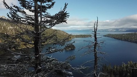 Drone flys to the left from behind a tree revealing Fannett Island and Emerald Bay Lake Tahoe California on a partly cloudy day some snow in the trees