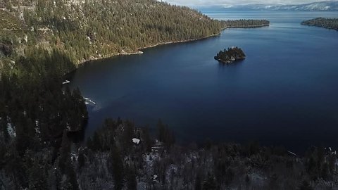 Drone flys from forest to water over emerald bay lake tahoe california revealing the bay. some snow on the beach and in the trees. partly cloudy day