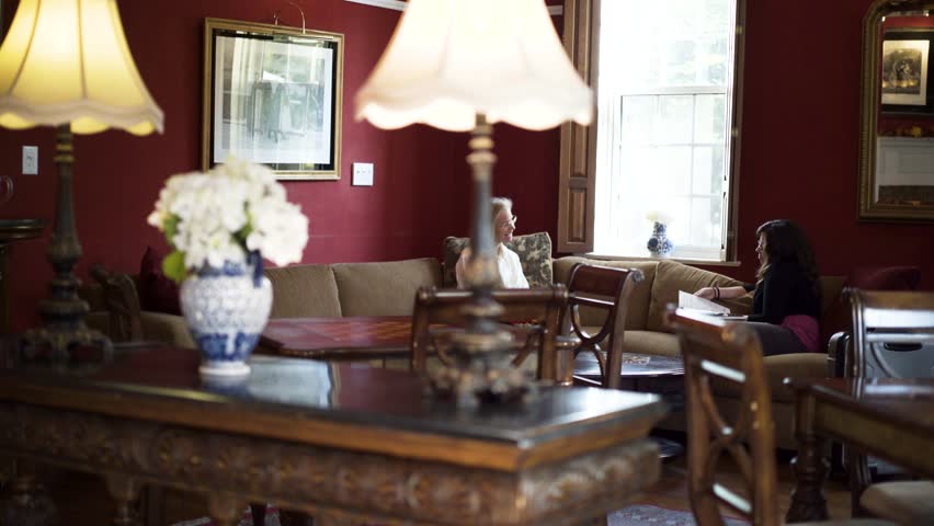 Two women sit on couches and talk in a grand room in a historic inn with red walls and large windows.